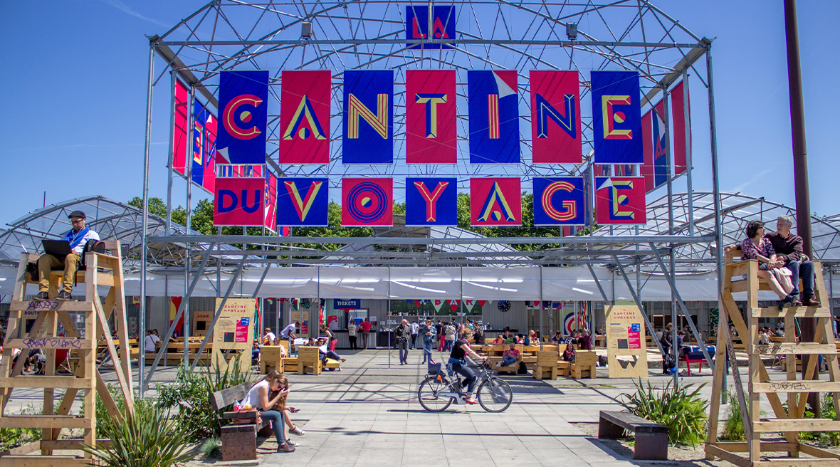 open plan restaurant with a large colourful sign saying la cantine du voyage with people sitting in the sun or riding bicycles past